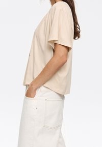 Beige short-sleeve top made from soft fabric, featuring a relaxed fit and subtle seam detailing, paired with light-colored high-waisted pants.