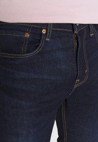 Tmavě modré džíny vyrobené z denimu s kontrastním žlutým prošíváním, disponující předn�ím zipem, zapínáním na knoflík a hlubokými kapsami. Hladká textura.
