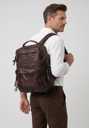 Man met een wit overhemd en een bruine broek draagt een grote donkerbruine leren rugzak over één schouder tegen een effen achtergrond.