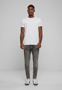 Camiseta de algodón blanca combinada con jeans ajustados grises claros y zapatillas blancas con detalles grises y un logo azul, de pie contra un fondo gris.