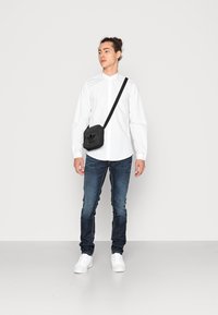 Camicia bianca a maniche lunghe, jeans in denim blu, borsa a tracolla nera con logo e sneakers bianche. Outifit semplice e casual.