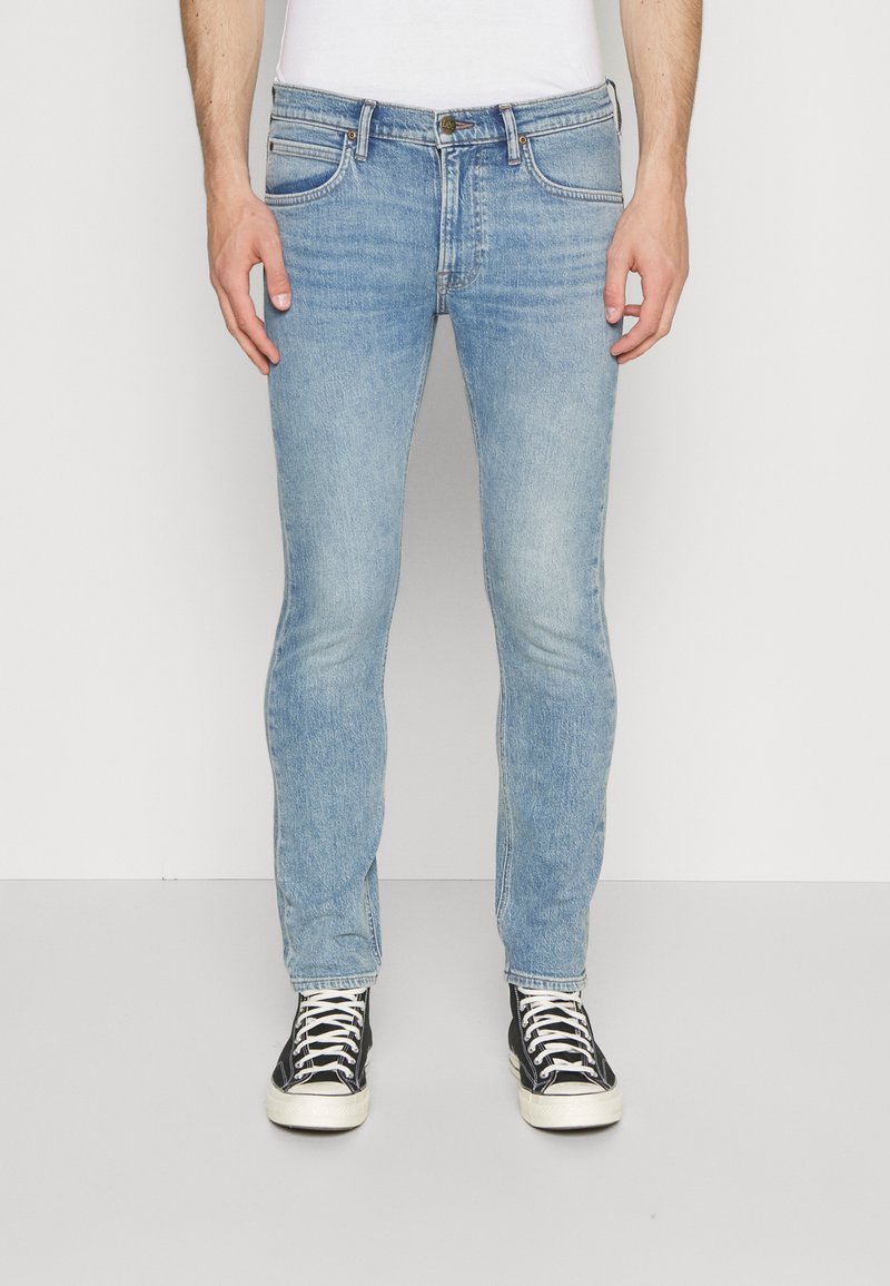 Jeans de mezclilla azul claro con corte ajustado, que presentan costuras en contraste, cinco bolsillos y un cierre de botón. Llevados con zapatillas negras y blancas.