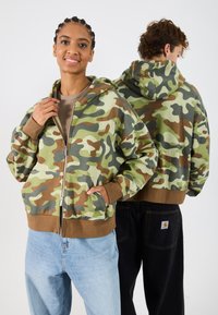 Sudadera con cremallera de camuflaje en tonos verdes, marrones y beiges; cuenta con capucha, puños acanalados y dobladillo, combinada con jeans de color azul claro.