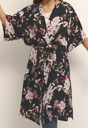 Kobieta nosząca czarny kwiecisty szlafrok w stylu kimono z szerokimi rękawami i wiązanym paskiem w talii, na białym koronkowym topie i dżinsach.