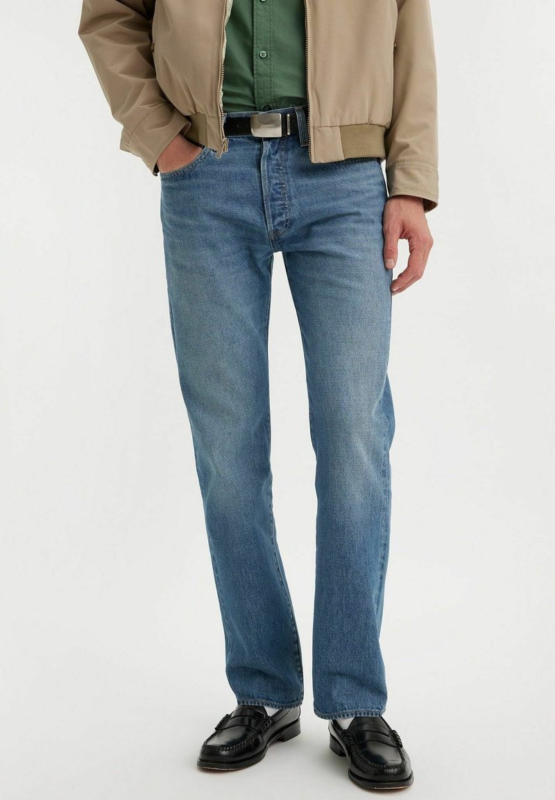 Homme portant un jean droit bleu clair, une ceinture noire, une chemise verte, une veste beige et des mocassins en cuir noir, debout devant un fond blanc.