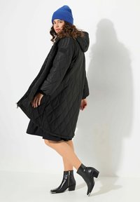 Cappotto lungo imbottito nero con cappuccio, dotato di cerniere laterali. Abbinato a un berretto blu a coste e stivaletti neri con tacco.