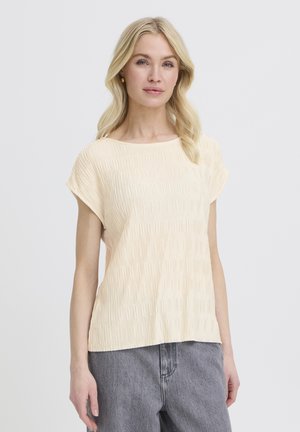 Blonde woman wearing a short-sleeve cream textured top and high-waisted gray jeans standing against a plain background.