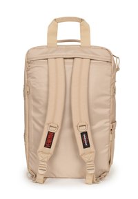 Beige ryggsäck med vadderade justerbara axelremmar, dubbla handtag, sidoficka med dragkedja och svarta logotypdetaljer. Slät nylonväv.