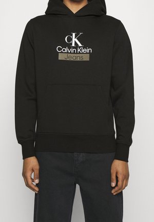 Czarna bluza z kapturem z logo "cK Calvin Klein Jeans" w białym i szarym kolorze na piersi, noszona przez osobę z rękami swobodnie opuszczonymi wzdłuż boków.