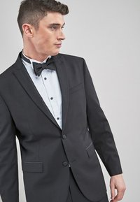 Schwarze Smokingjacke mit satinierter Revers, einknöpfigem Verschluss und Seitentasche, kombiniert mit einem weißen Hemd und einer schwarzen Fliege.