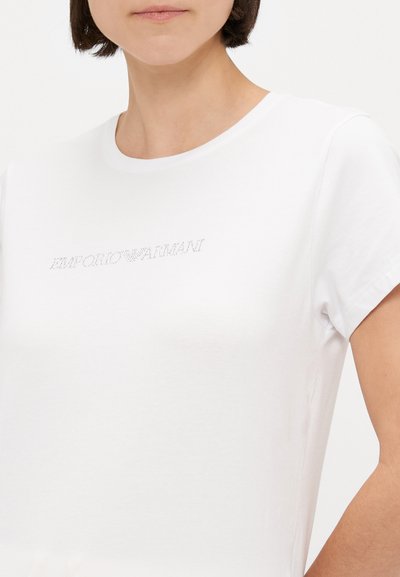 Bjela pamučna majica s okruglim izrezom, kratkim rukavima i logotipom ukrašenim rhinestone-om na prsima. Glatka tekstura.