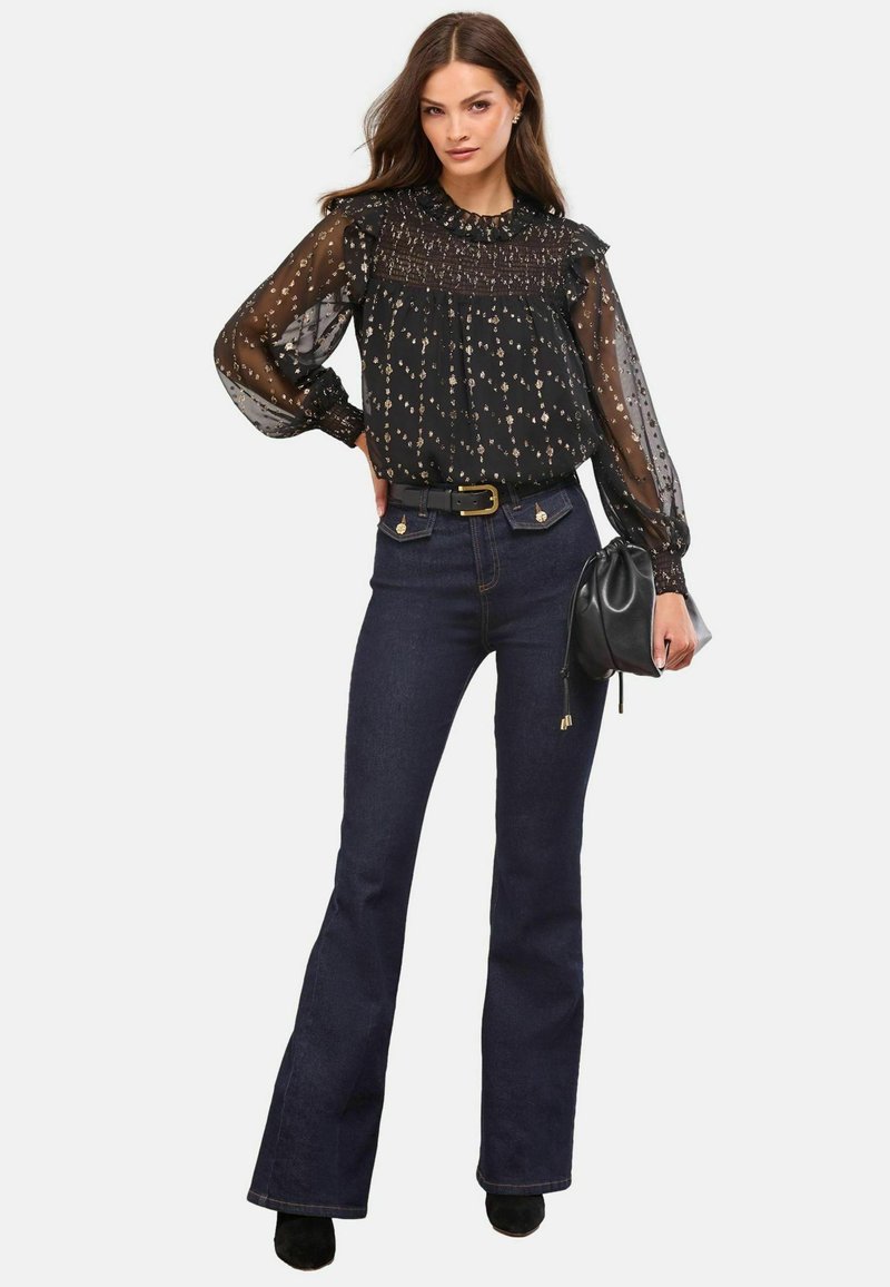 Schwarze durchsichtige Bluse mit floralem Muster und Rüschenausschnitt, kombiniert mit dunkelblauen ausgestellten Jeans und einer schwarzen Handtasche, getragen mit schwarzen Schuhen.