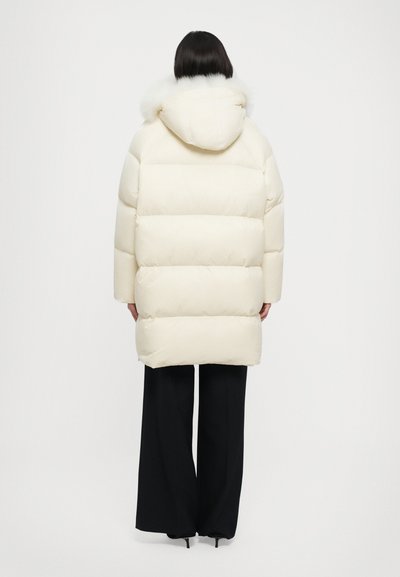 Pufferschale in Creme mit einer Kapuze aus Kunstfell, kastigem Design und horizontaler Steppung. Kombiniert mit weit geschnittenen schwarzen Hosen.