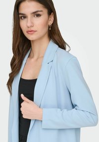 Gaiši zils žakete ar izgriezumiem atloks, teksturētu audumu un garām piedurknēm, vilkts pār melnu topu. Modele pozē ar roku uz gurna.