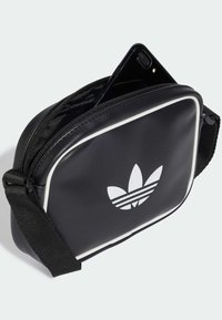 Čierna syntetická kožená taška s bielym prešívaním, na ktorej sa nachádza výrazné logo Adidas trefoil. Obsahuje zips a nastaviteľný popruh.