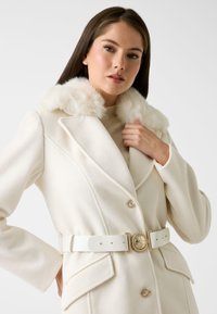 Weißer Wollmantel mit einer flauschigen Kunstfellkapuze, doppelreihigem Design und einem breiten weißen Gürtel mit einer goldenen, runden Schnalle. Texturiertes Material.