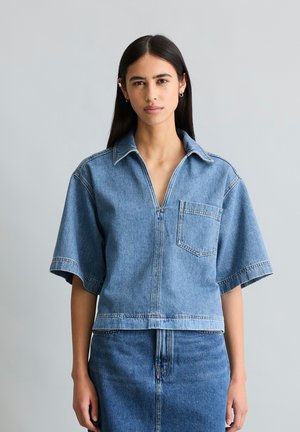 Femme portant une chemise en jean bleue à manches courtes avec col et poche avant, associée à une jupe en jean assortie, debout devant un fond gris.