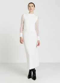 Langes weißes Kleid mit taillierter Korsage, strukturierten Spitzen-Longärmeln und gerippten Bündchen; kombiniert mit schwarzen Stilettos mit Knöchelriemen.