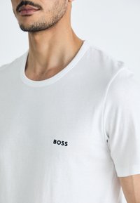T-shirt en coton blanc avec un col rond et des manches courtes, arborant un logo "BOSS" noir brodé sur la poitrine gauche.