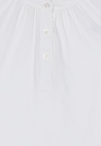 Närbild av vitt tyg som visar en skjortknäppning med fyra små vita knappar och subtila veck upptill.