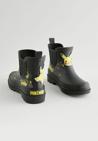 Bottes en caoutchouc noires à la cheville avec une finition texturée, arborant des graphismes jaunes de Pikachu et des motifs de projection, ainsi qu'un logo "Pokémon" bien en vue sur le talon.