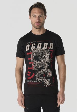 Hombre vistiendo camiseta negra con gráfico de dragón blanco y rojo y texto que incluye "Osaka" y caracteres japoneses, de pie frente a un fondo liso.