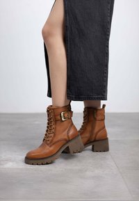 Botas de tobillo de cuero marrón con cordones en la parte delantera, hebilla decorativa y tacón de goma grueso. Superficie texturizada y cremallera lateral para facilitar su uso.