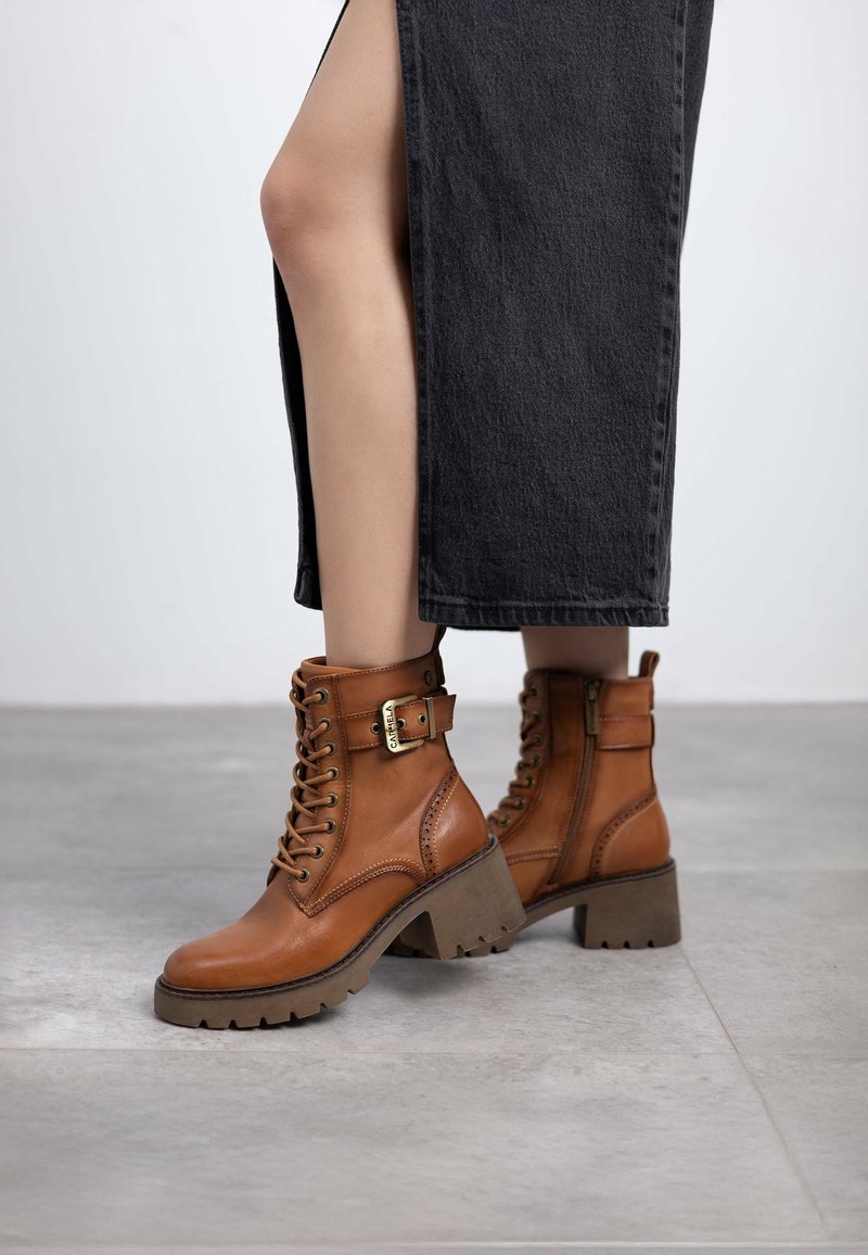 Botas de tobillo de cuero marrón con cordones en la parte delantera, hebilla decorativa y tacón de goma grueso. Superficie texturizada y cremallera lateral para facilitar su uso.