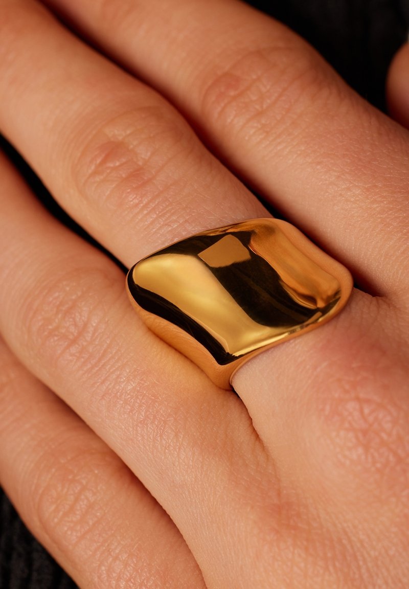 Gouden, dikke ring met een gladde, gepolijste oppervlakte, met een afgeronde vorm en lichte asymmetrie, gedragen aan een hand met een gestructureerde huid.