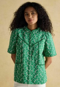 Blusa verde a maniche corte con intricato motivo in pizzo floreale bianco, rifiniture decorative e chiusura con bottoni davanti. Tessuto morbido.