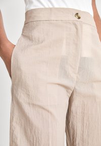 Pantaloni in tessuto leggero beige, con texture stropicciata, caratterizzati da una chiusura frontale con bottone e tasche laterali, progettati per una vestibilità comoda.