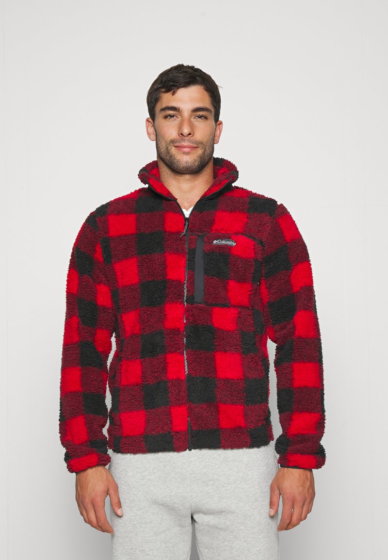 Veste en polaire à carreaux rouges et noirs, avec un col zippé, une poche poitrine noire et une finition douce et texturée. Un pantalon de survêtement gris est porté en dessous.