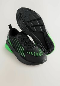 Chaussures de sport noires avec des accents verts, tige en maille texturée, semelle en caoutchouc et motifs sur les côtés et la semelle pour une meilleure adhérence.