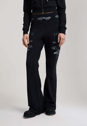 Kobieta nosząca czarne, rozszerzane spodnie z białymi wzorami pazurów i napisem "Ed Hardy" na pasie, w połączeniu z czarnymi butami i czarnym topem.
