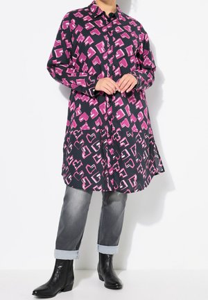 Person iført en lang sort skjorte med pink abstrakte mønstre, op rullede grå jeans og sorte ankelstøvler, stående mod en hvid baggrund.