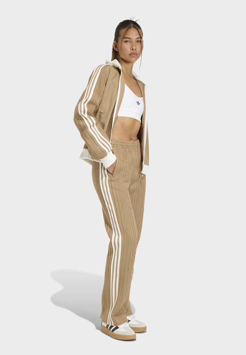 Tan athletic tracksuit with ribbed texture, featuring white three-stripes on sleeves and pants. White crop top and sneakers with black accents.