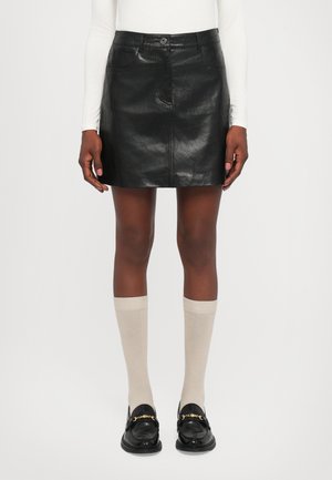 Vrouw in een zwart leren mini-jurk, beige kniekousen, zwarte loafers met gouden ketting en een witte langemouwenshirt, staand tegen een witte achtergrond.