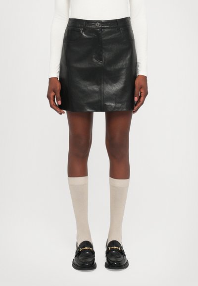 Kvinde iført en sort læder miniskørt, beige knæhøje sokker, sorte loafers med guld kæde og en hvid langærmet top, stående mod hvid baggrund.
