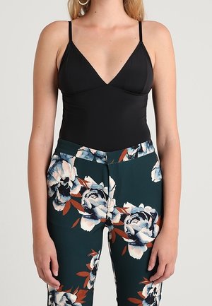 Vrouw draagt een zwarte mouwloze top met V-hals en donkere bloemenbroek met grote witte en oranje bloemen, staand met ontspannen armen.