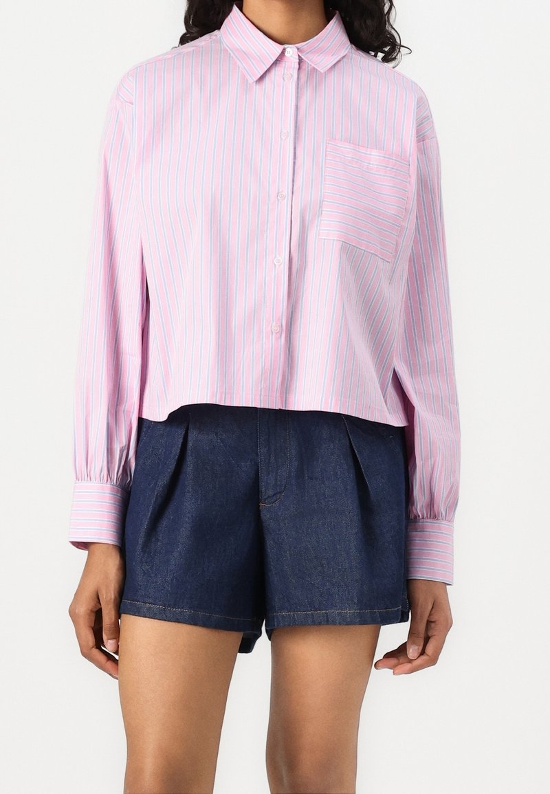 Femme portant une chemise à boutons rayée rose clair et blanche avec manches longues et un short taille haute bleu foncé.