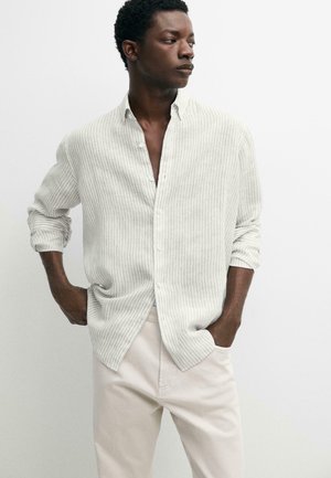 Uomo che indossa una camicia beige chiaro a righe con bottoni e maniche rimboccate e pantaloni color crema, mani in tasca, guarda di lato.