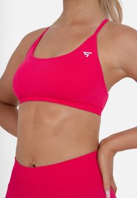 Reggiseno sportivo rosa con spalline sottili, realizzato in tessuto morbido e elastico. Presenta un piccolo logo bianco sul lato sinistro.