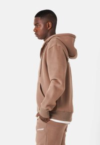 Felpa con cappuccio in cotone marrone, con tasca frontale, polsini e orlo a coste. Indossata con pantaloni abbinati, con un logo sul petto.