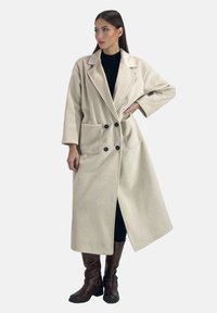 Beige, lång, dubbelknäppt kappa med stora fickor och svarta knappar. Tillverkad av mjukt tyg, med kavajkrage och vida ärmar.