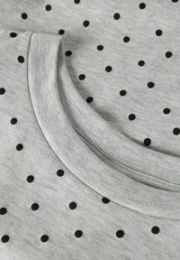 Grauer Stoff mit einer weichen Textur und einem schwarzen Tupfenmuster. Enthält einen abgerundeten, gerippten Kragen.