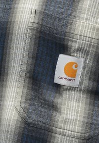 Plaid marškiniai, kuriuose vyrauja pilka, mėlyna ir balta spalvos. Pagaminti iš tekstūruoto audinio, turi krūtinės kišenę ir Carhartt logotipų etiketę oranžinės ir baltos spalvos.