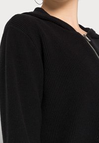 Sweat à capuche noir à fermeture éclair avec un tissu texturé côtelé. Comprend une capuche ajustée et des détails de fermeture éclair argentés à l'avant.