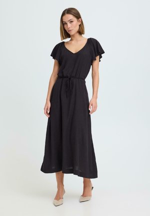 Kvinde iført sort midi-kjole med flagrende ærmer og bindebånd i taljen, stående mod en ensfarvet lys baggrund iført beige spidse flade sko.