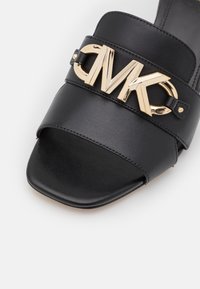 Black leather slide sandal with a wide strap featuring a gold MK logo. Square toe design with stitching details on the upper.