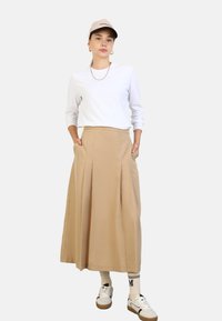Gonna midi plissettata beige con due tasche laterali, abbinata a una maglietta bianca a maniche lunghe e scarpe da ginnastica bianche con dettagli beige.